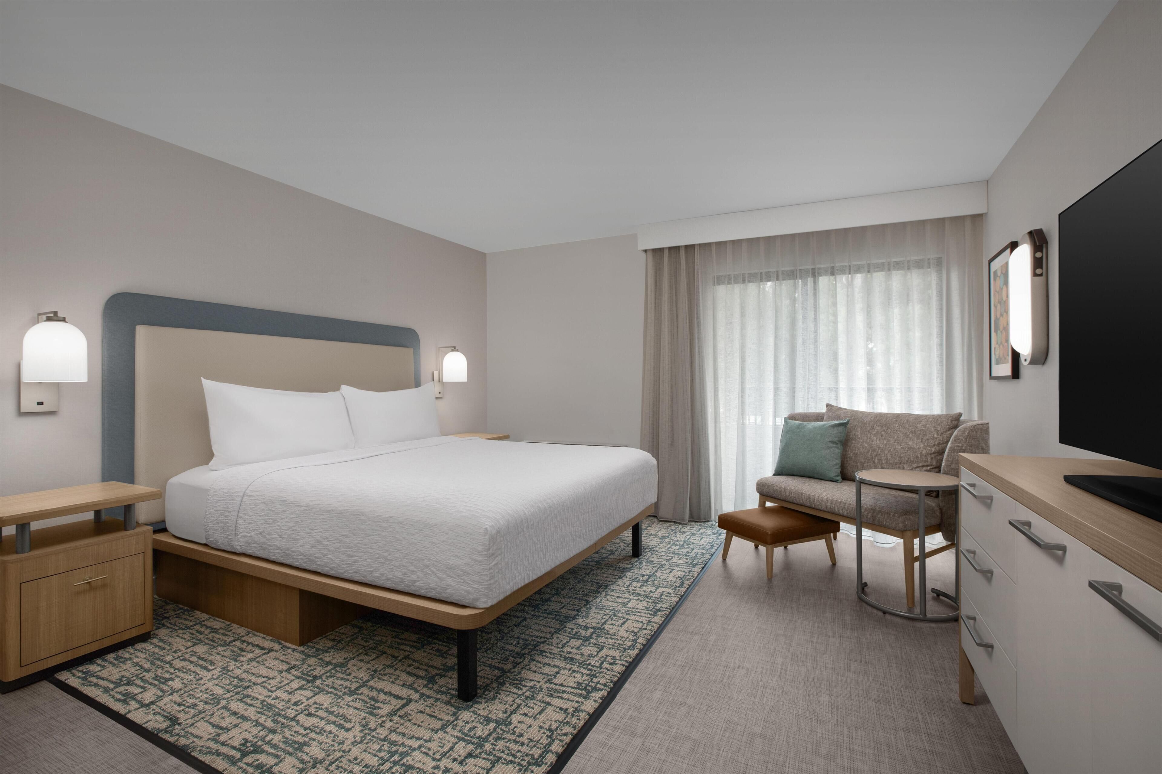 A hotel room with a bed, bedside tables, a desk with a chair, a television, and a large window with curtains.