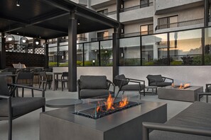 Un espacio de estar al aire libre con mobiliario moderno, una chimenea con cubierta de vidrio y vista a un edificio con balcones.