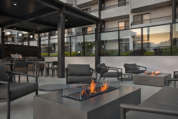 Un espacio de estar al aire libre con mobiliario moderno, una chimenea con cubierta de vidrio y vista a un edificio con balcones.