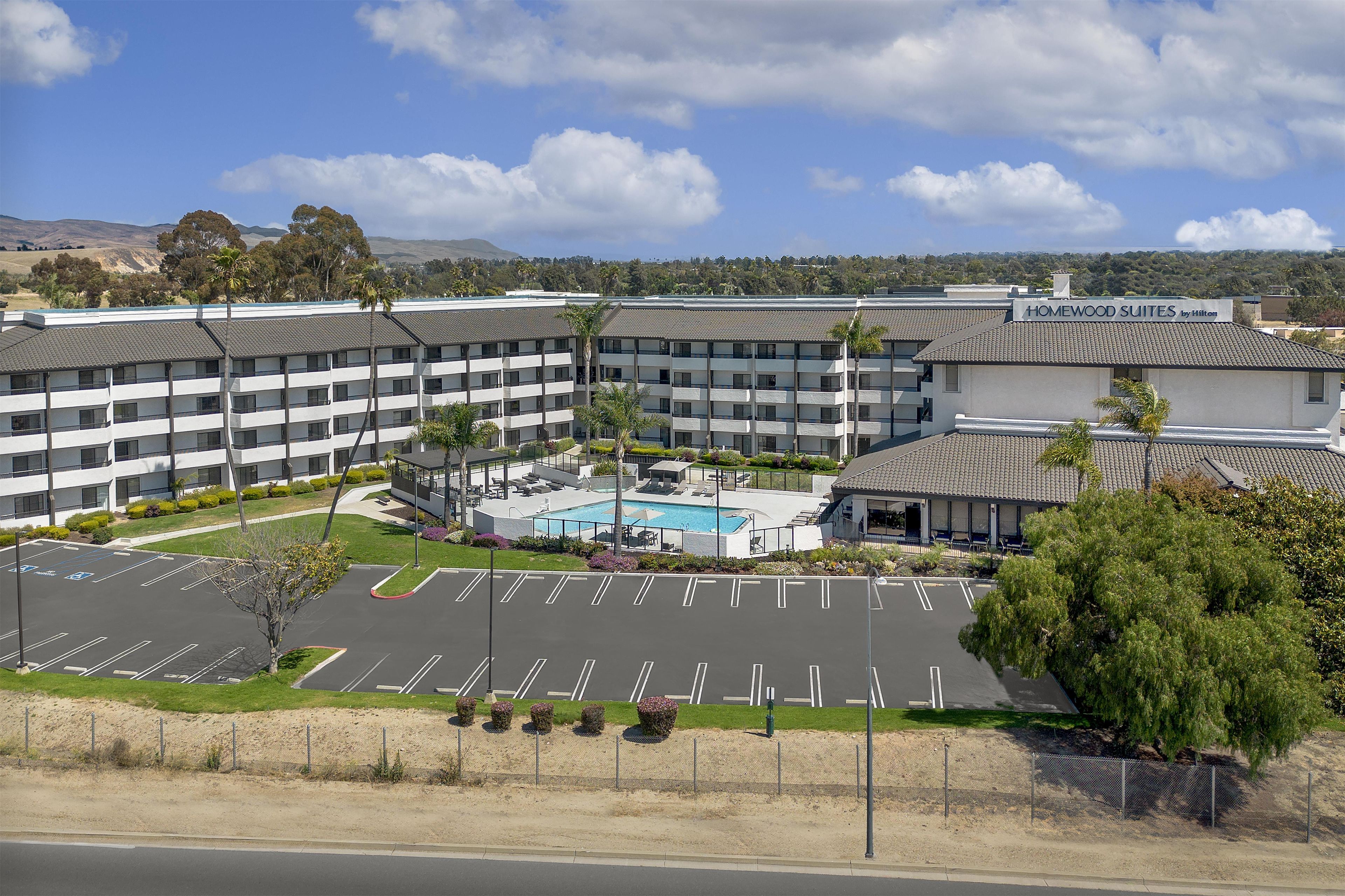 A multi-story hotel with a swimming pool, parking lot, and a view of the surrounding landscape.