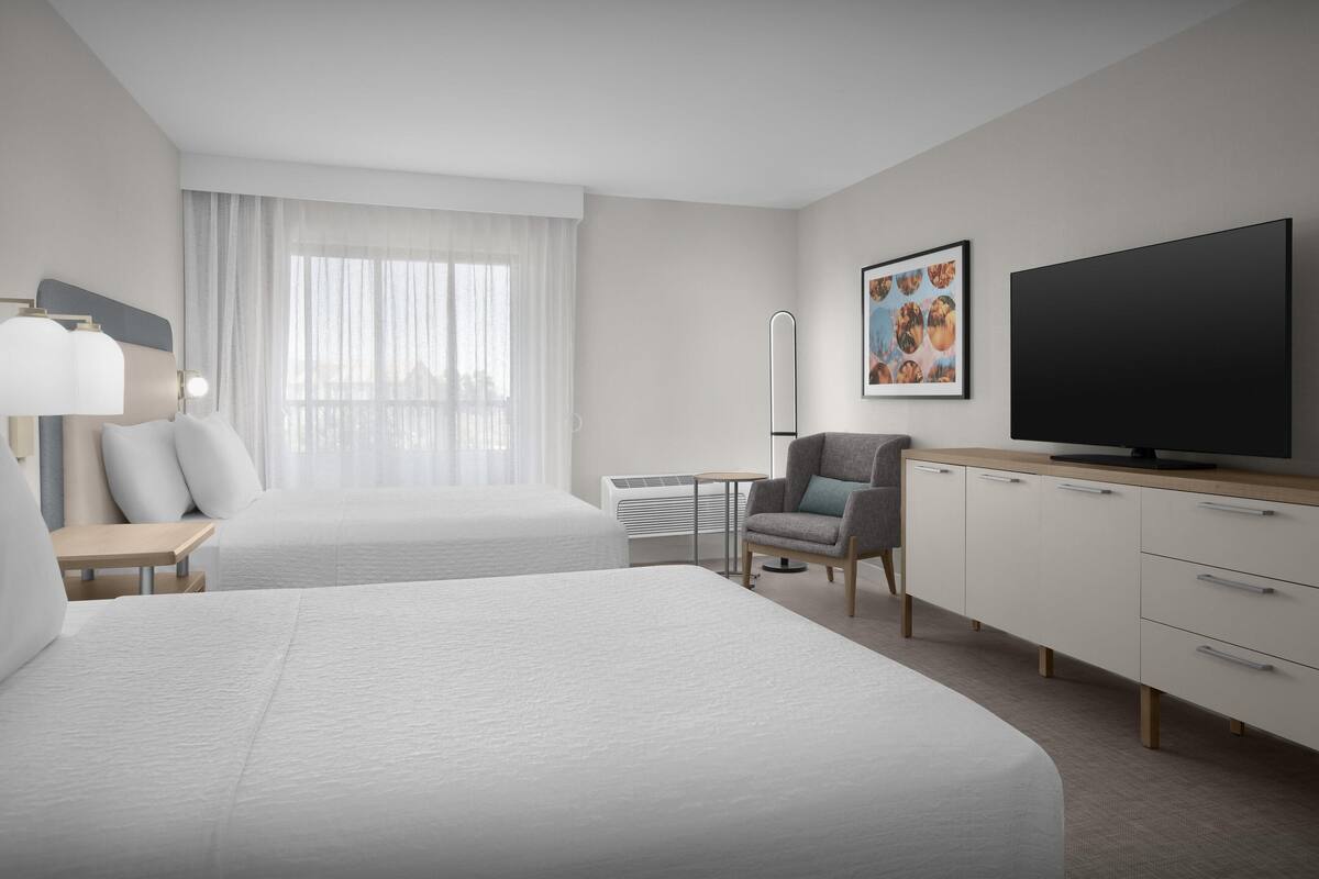 A hotel room with two beds, a TV, a chair, a desk, and a window with sheer curtains.