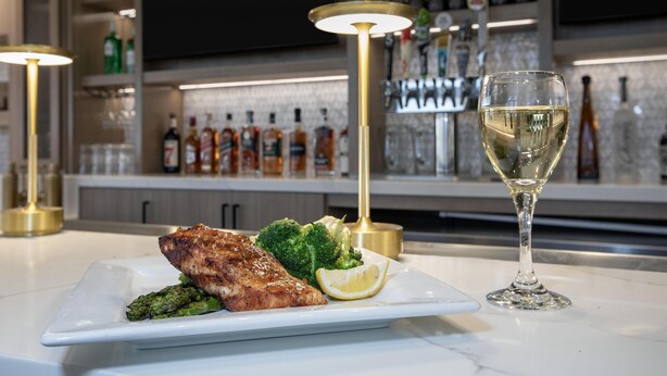 Una mesa de comedor con un plato de comida, una copa de vino blanco y una zona de bar al fondo.