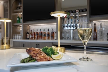 A dining table with a plate of food, a glass of white wine, and a bar area in the background.