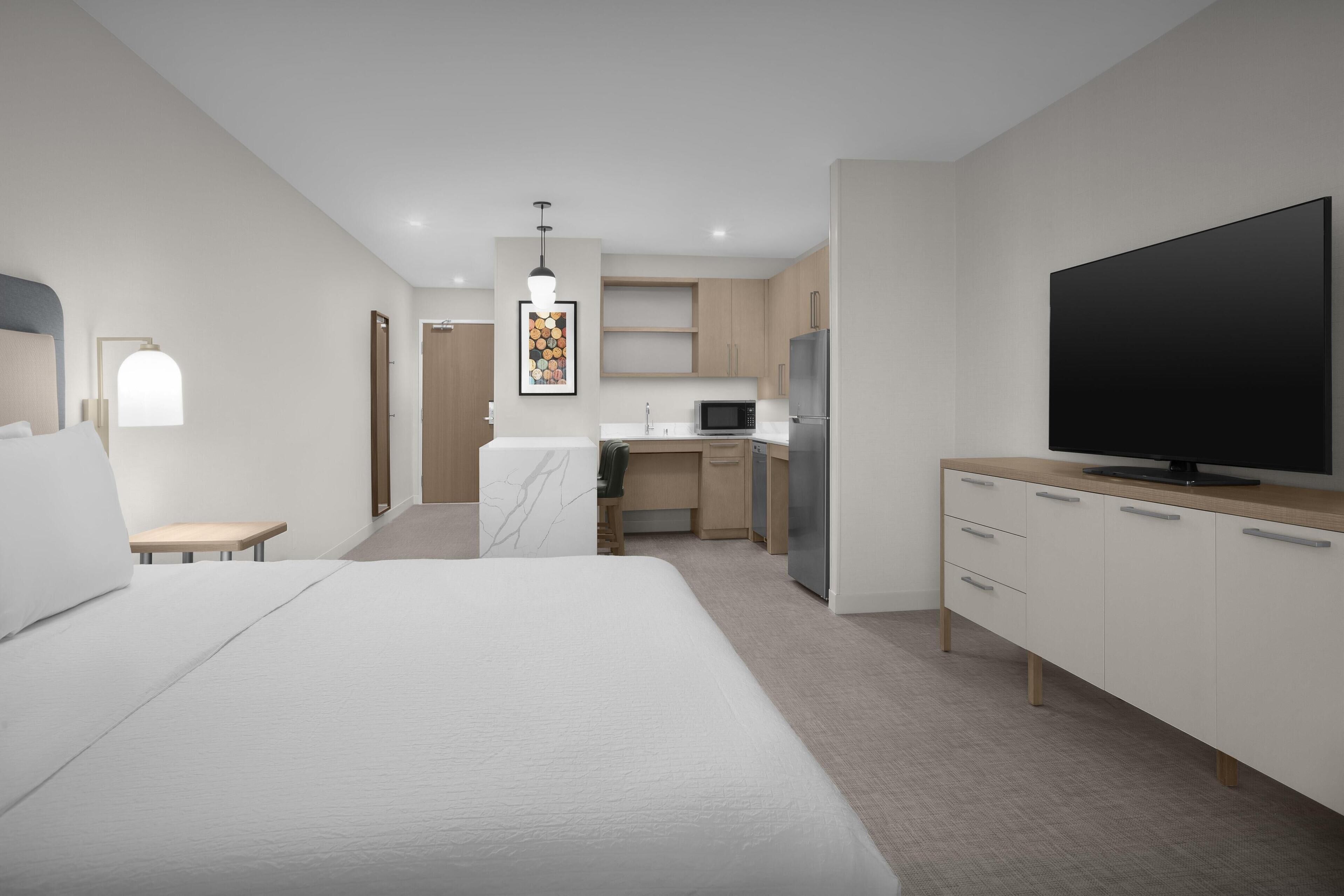 Una habitación de hotel moderna con una cama grande, televisión, zona de cocina y un pequeño rincón de descanso.