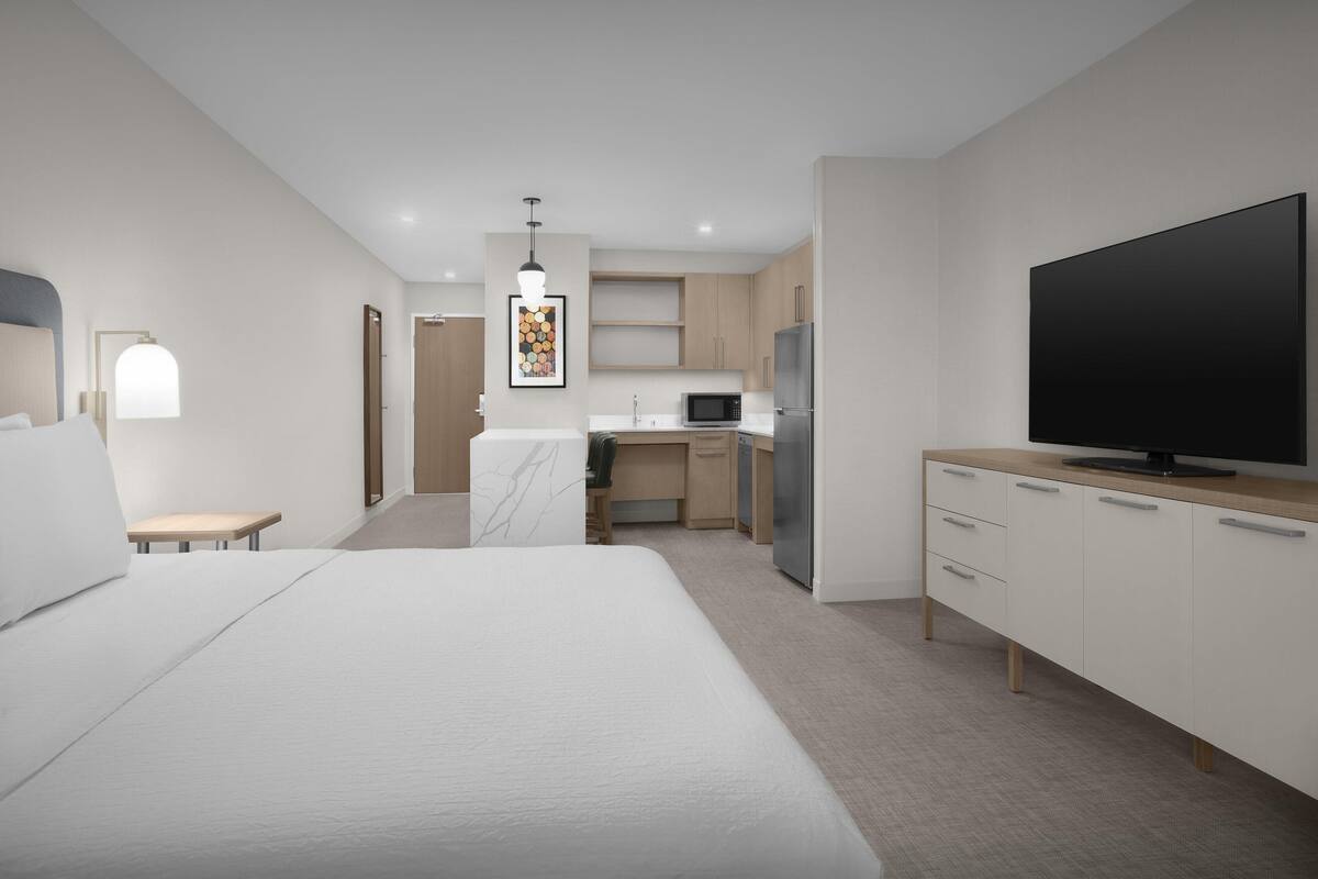 A modern hotel room with a large bed, a TV, a kitchen area, and a small seating area.