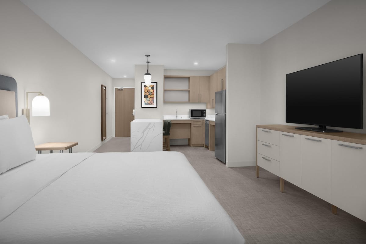 Una habitación de hotel moderna con una cama grande, televisión, zona de cocina y un pequeño rincón de descanso.