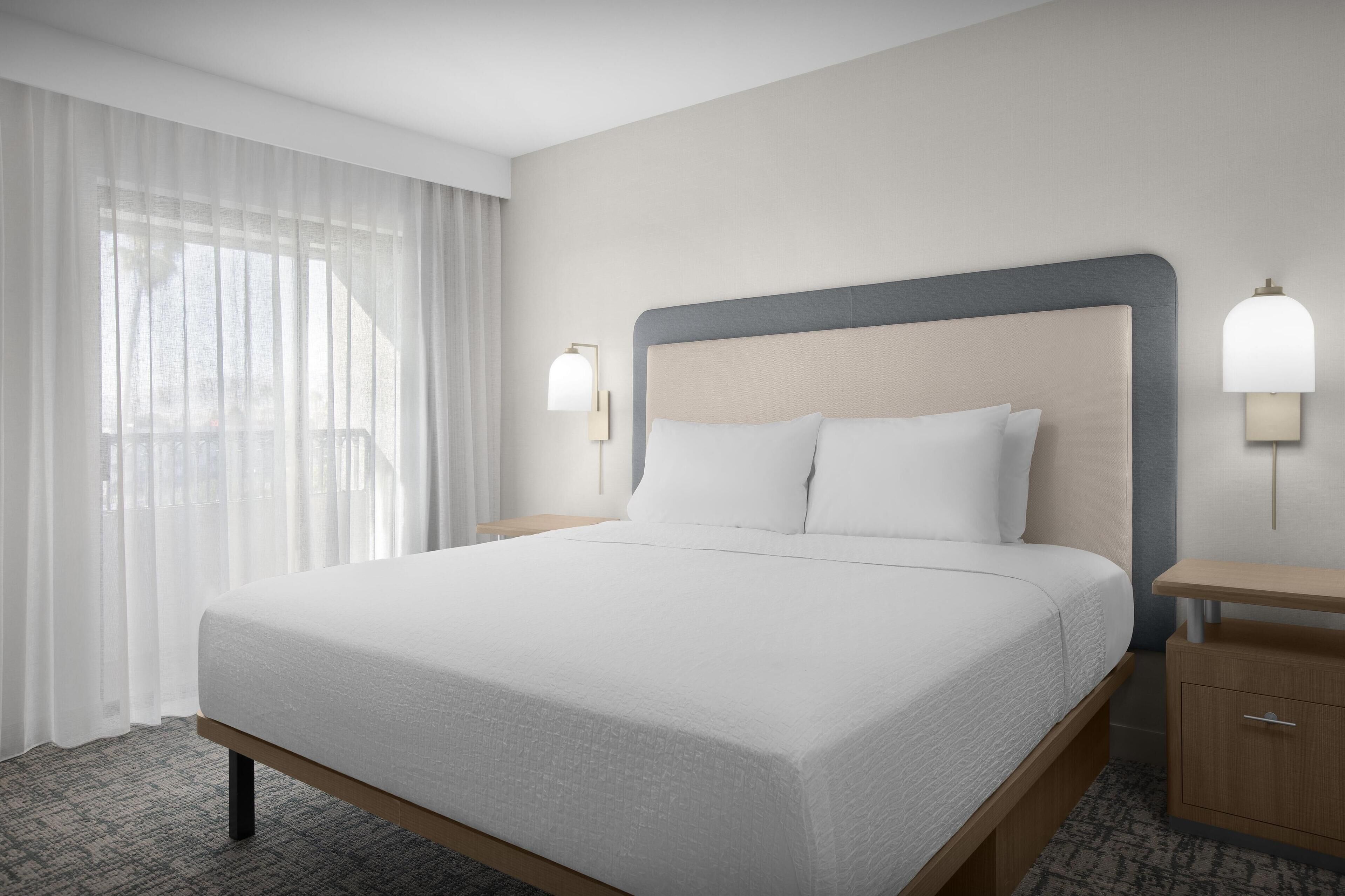 Una habitación de hotel con una cama grande, dos mesitas de noche y un ventanal con cortinas translúcidas.
