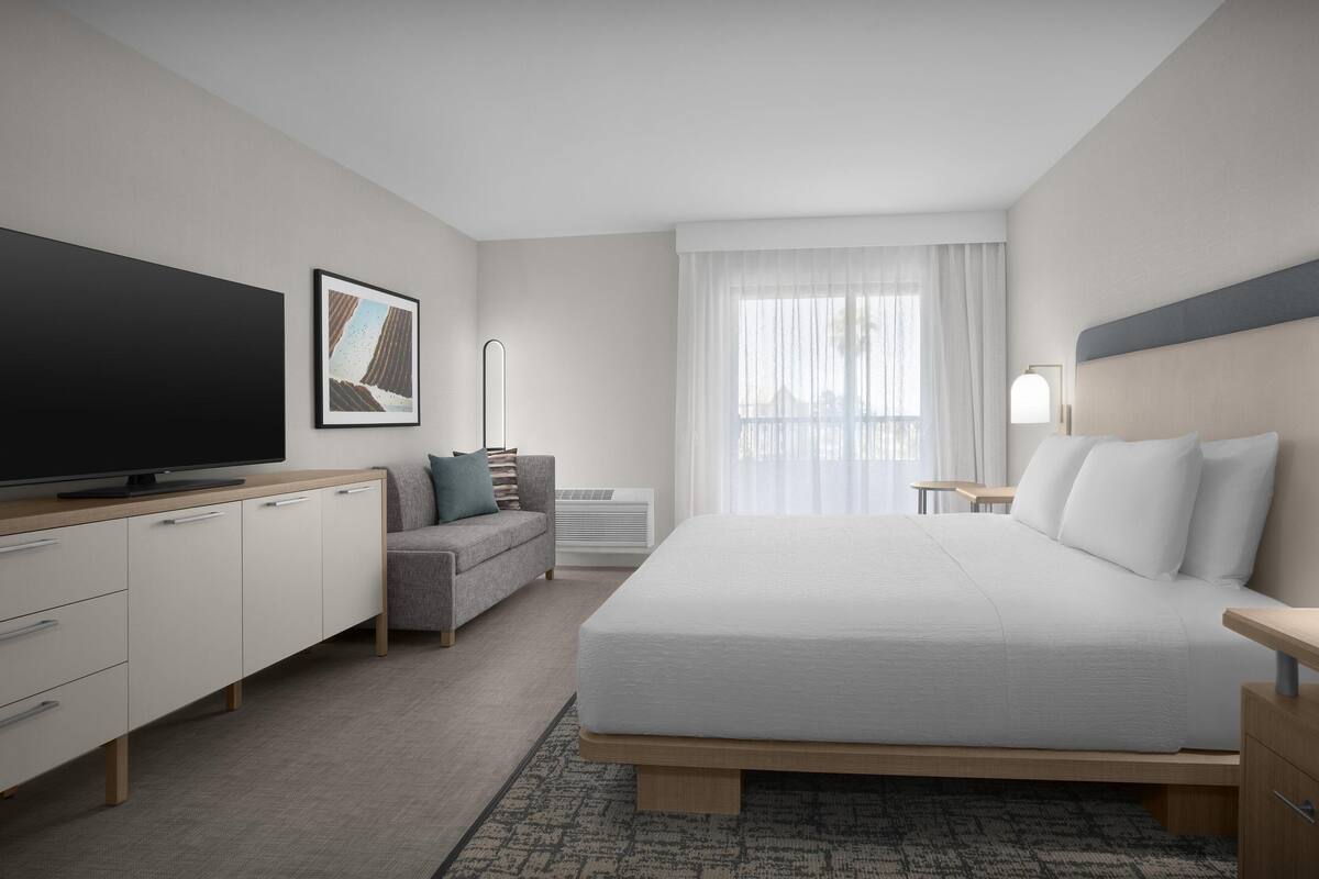 A modern hotel room with a bed, a TV, a sofa, a desk, and a window with curtains.