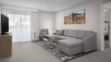 Ein modernes Wohnzimmer mit einem grauen Sofa, einem kleinen Tisch, einem Fernseher auf einem Holzschrank und einem großen Gemälde an der Wand.