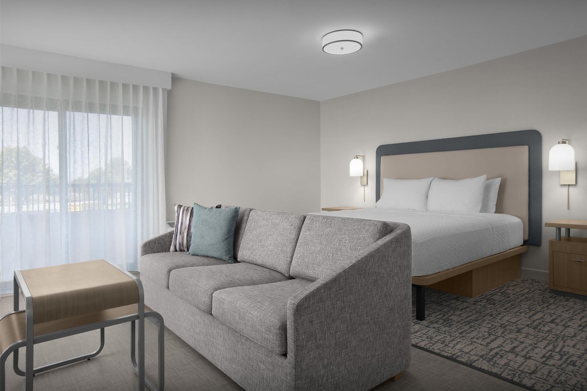 A modern hotel room with a grey sofa, a small wooden table, a bed with a grey headboard, bedside lamps, and a view through sheer curtains.