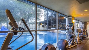 A gym with exercise equipment and a view of a swimming pool through large windows. - Hilton Dallas Lincoln Centre by the Galleria (Dallas)
