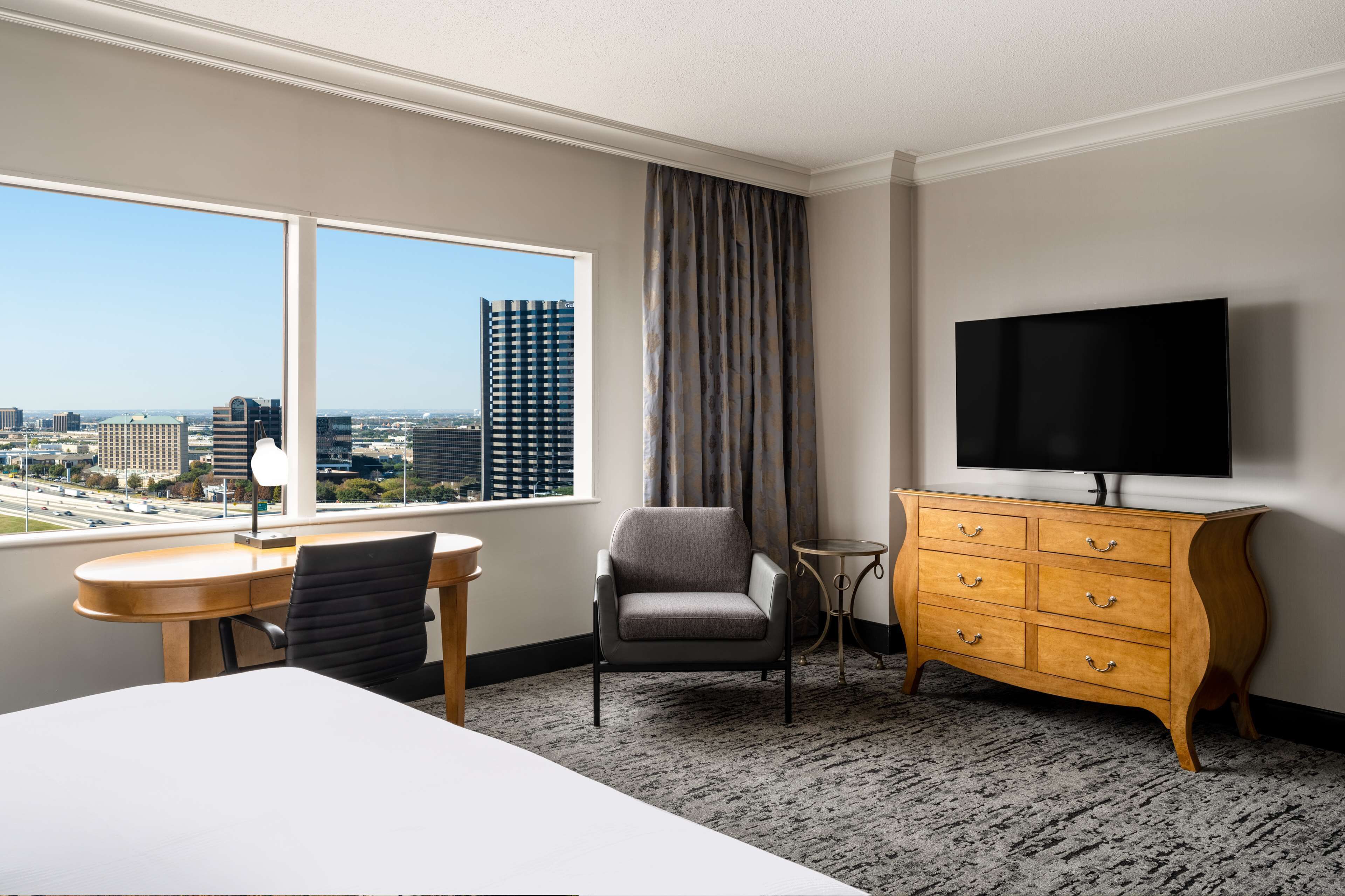 Une chambre d’hôtel avec une grande fenêtre, un bureau, une chaise, un téléviseur et une commode en bois.