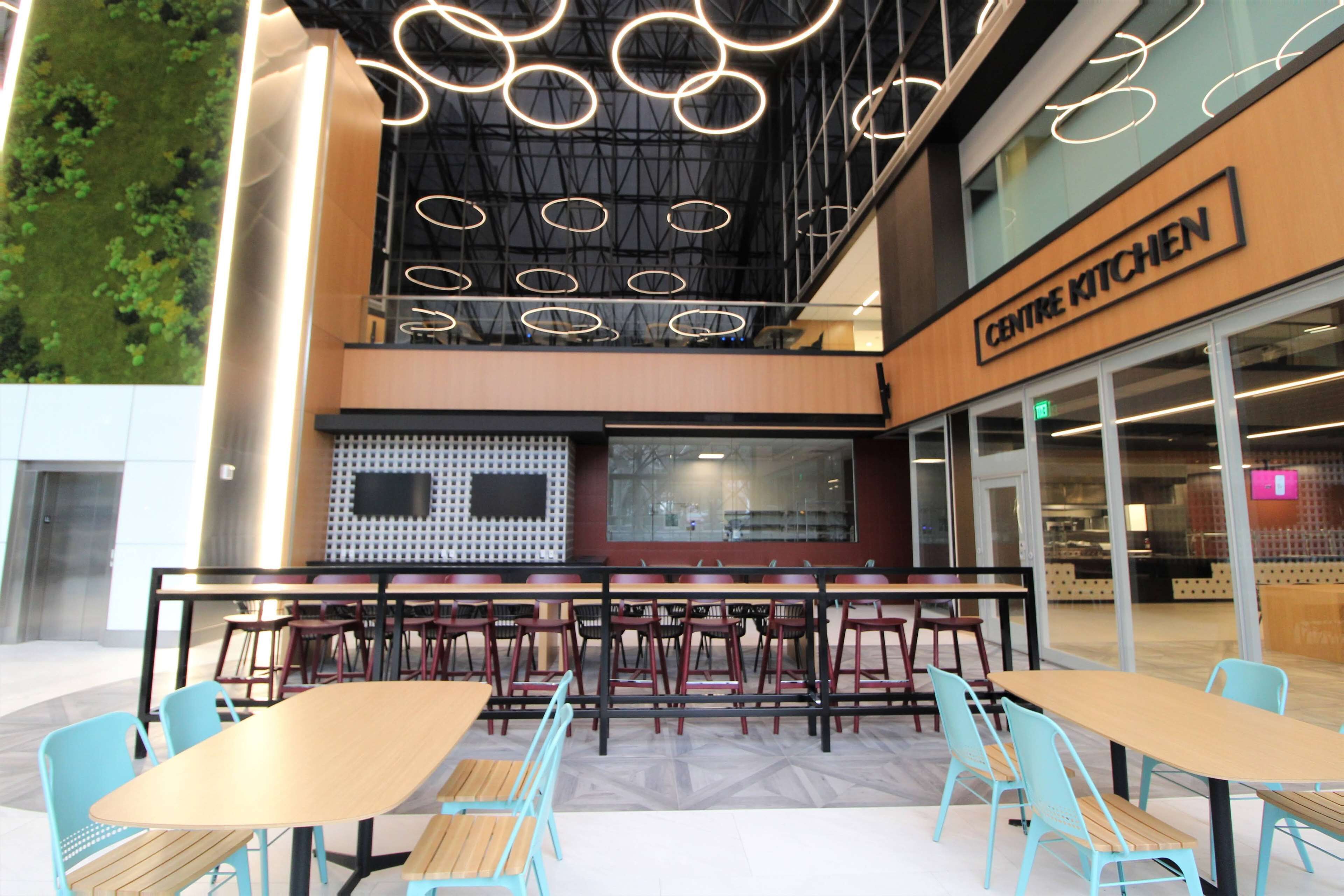 An outdoor seating area with tables and chairs, a sign for 'Centre Kitchen', and a modern interior design with a high ceiling and circular light fixtures.