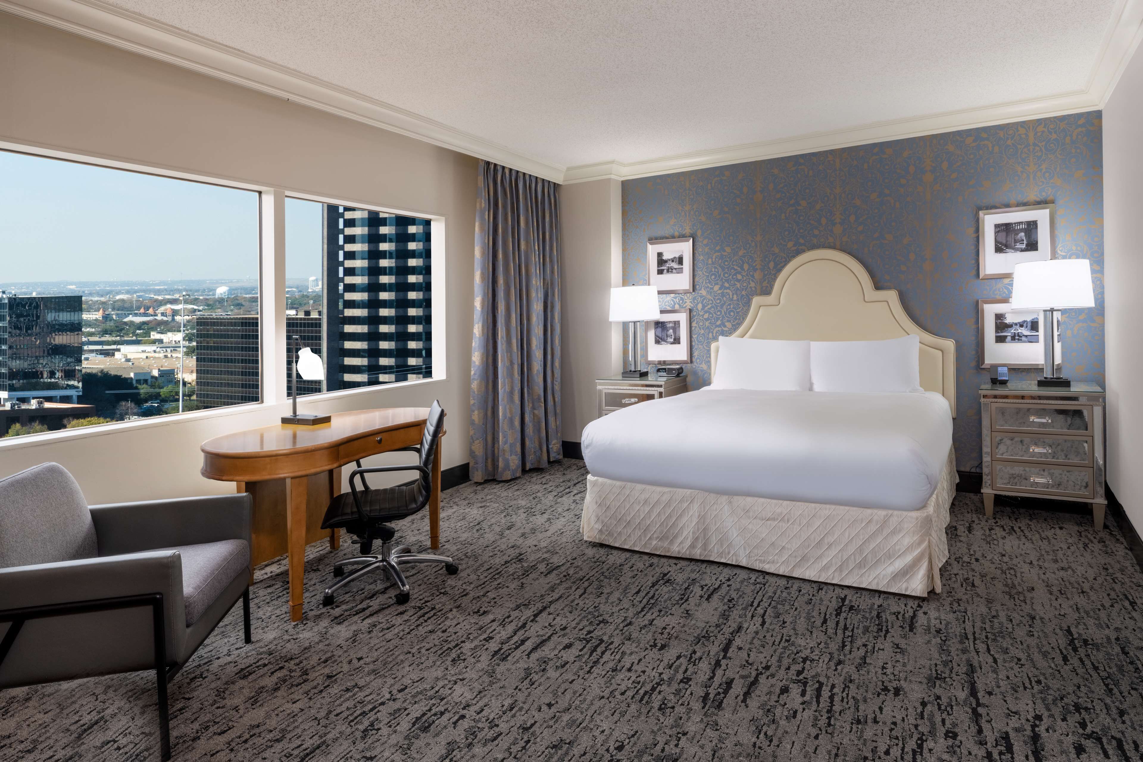 Ein Hotelzimmer mit einem großen Bett, einem Schreibtisch, einem Sessel und Blick auf die Skyline durch große Fenster.