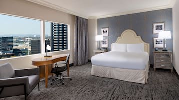 Ein Hotelzimmer mit einem großen Bett, einem Schreibtisch, einem Sessel und Blick auf die Skyline durch große Fenster.