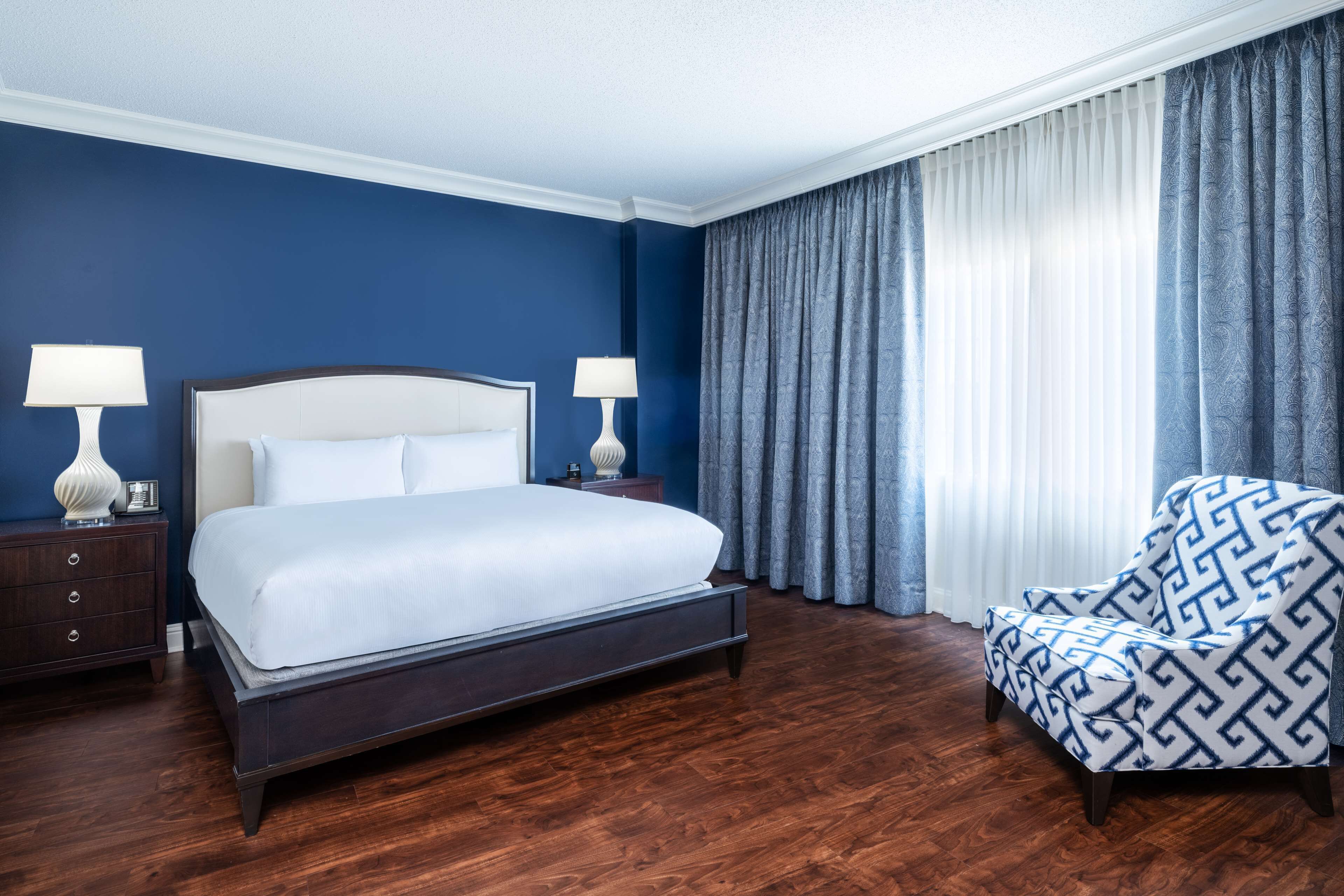 Een hotelkamer met een groot bed, twee nachtkastjes met lampen, een gestreept fauteuil en een raam met gordijnen.