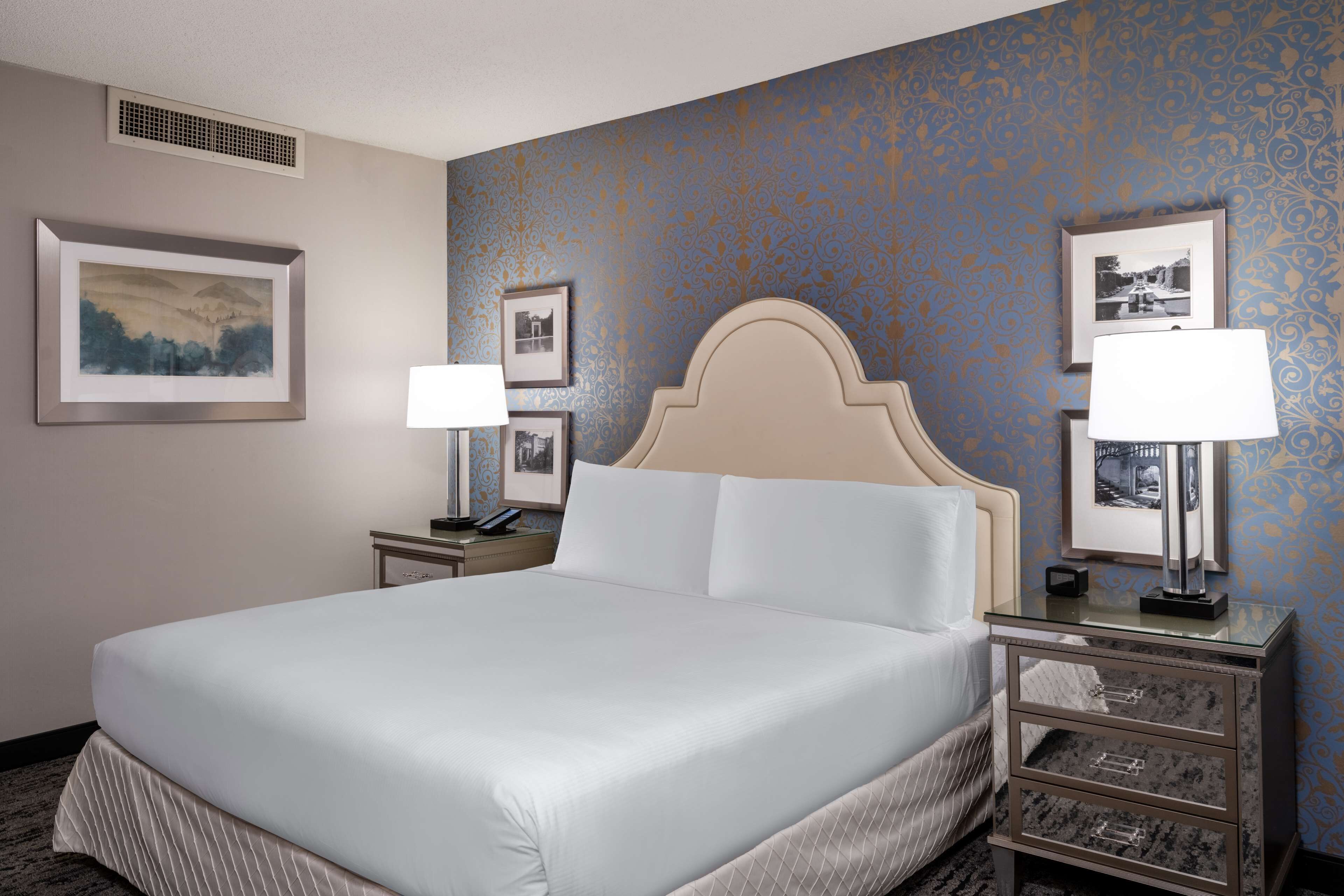 Una habitación de hotel con una cama grande, dos mesitas de noche con lámparas, un cabecero y cuadros enmarcados en las paredes.