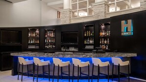 A bar area with a black countertop, white bar stools, and shelves of liquor bottles. - Hilton Dallas Lincoln Centre by the Galleria (Dallas)