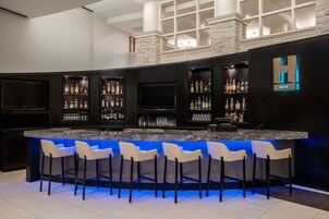 Uma área de bar com bancada preta, banquetas brancas e prateleiras com garrafas de bebidas.