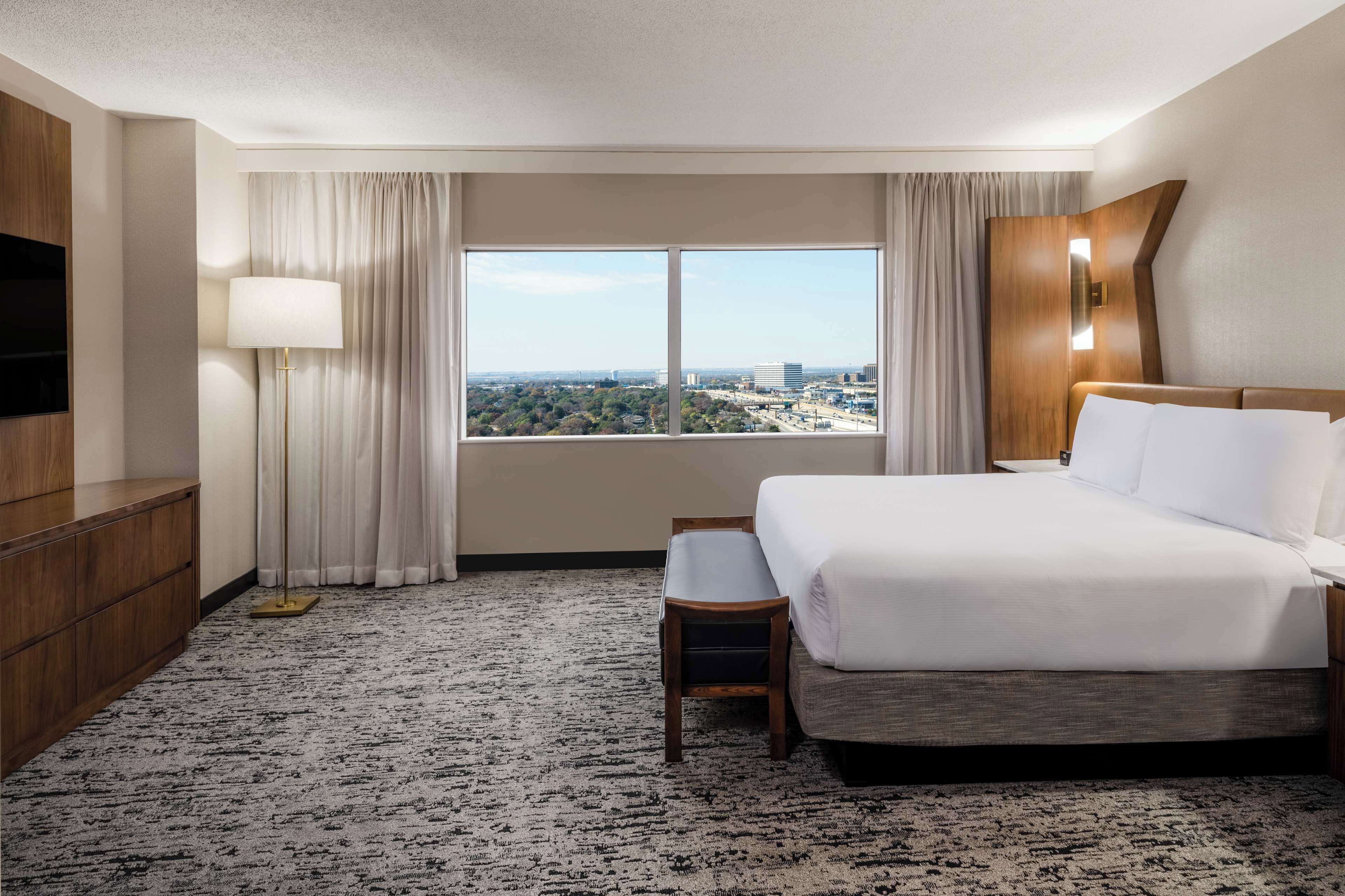 A hotel room with a bed, a desk, a chair, a TV, and a window with a city view.