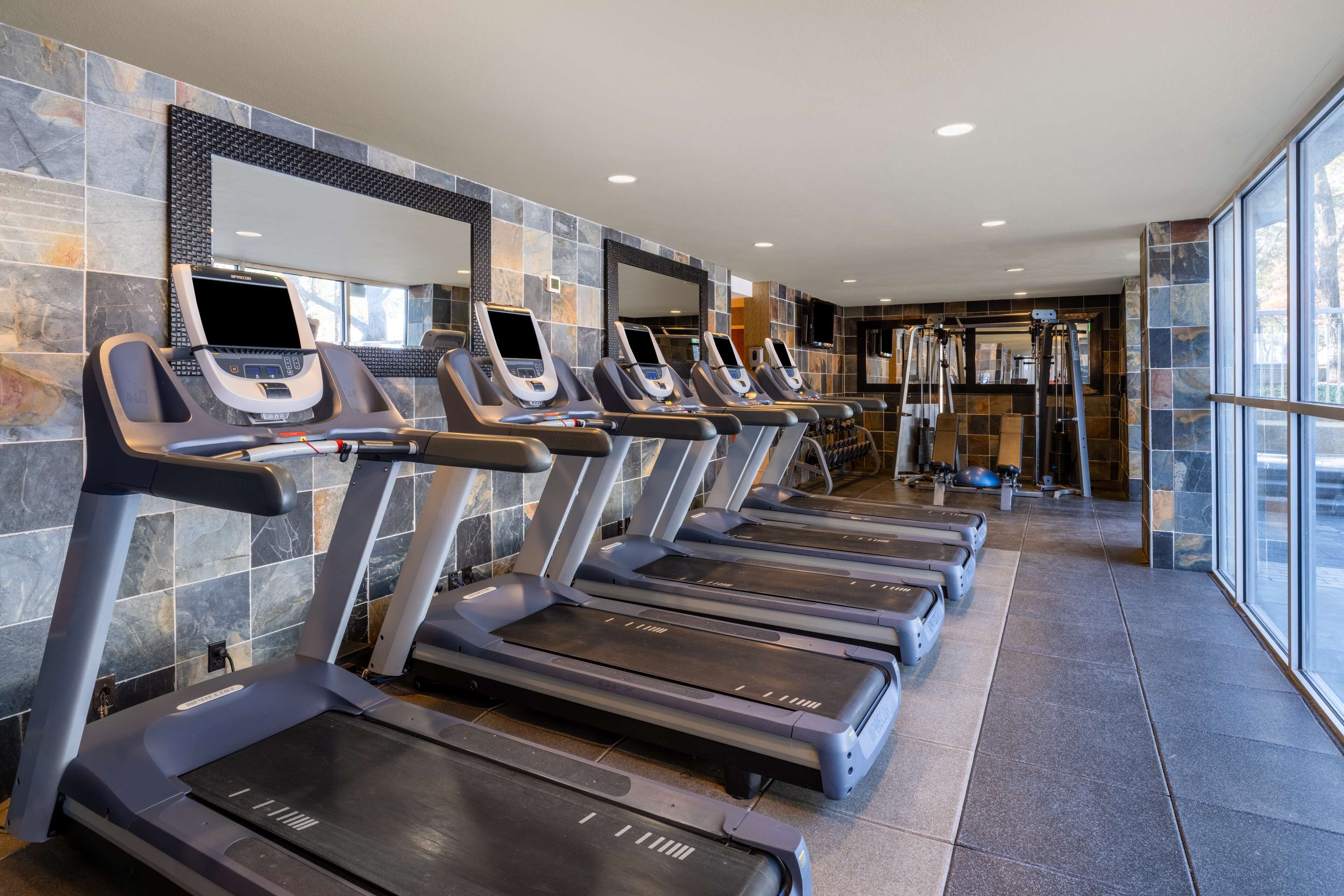 A row of treadmills with digital screens, a mirrored wall, and a stone-tiled floor.