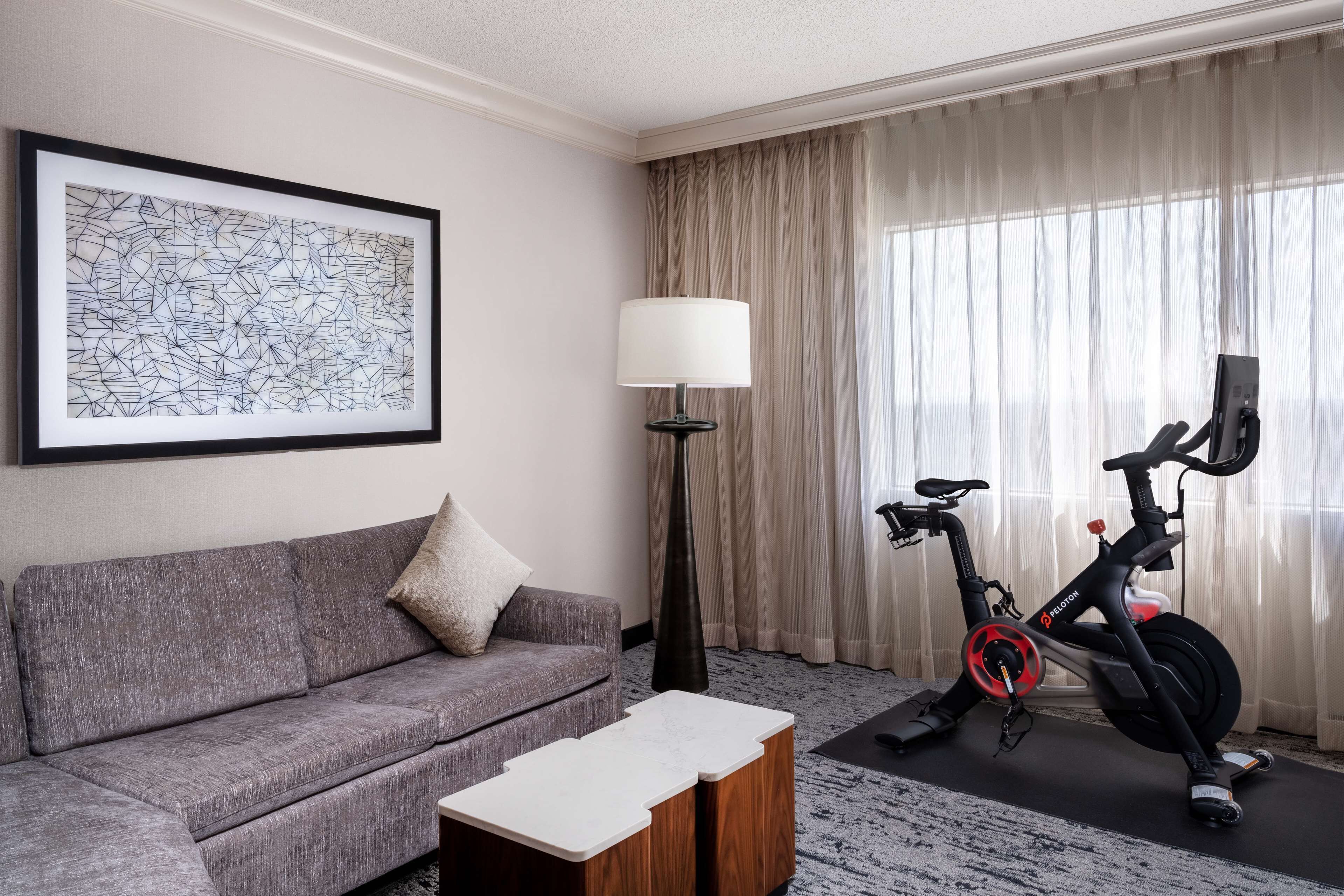 A modern hotel room with a grey sofa, a framed abstract artwork, a standing lamp, and a stationary exercise bike.