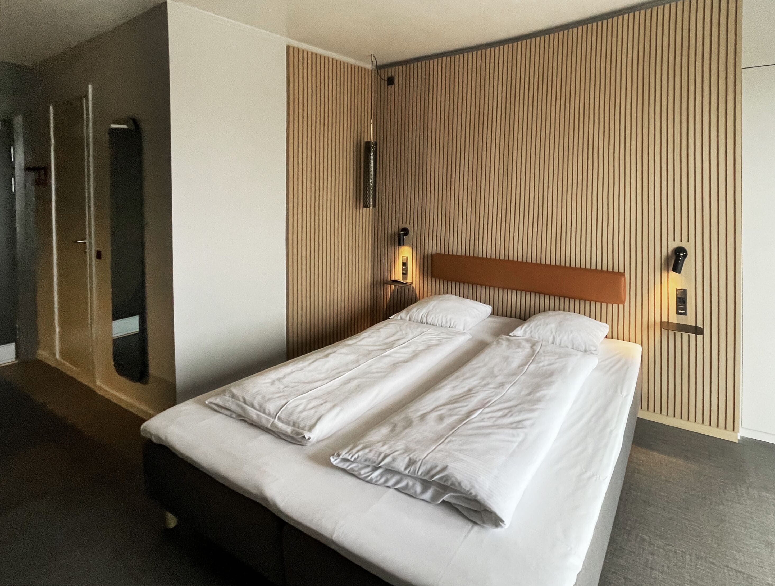 Una habitación de hotel con cama, mesita de noche, lámpara en la pared y una puerta.