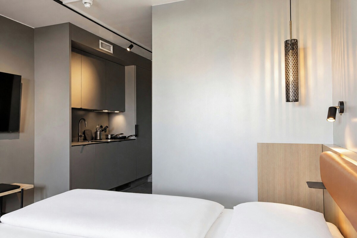 A modern hotel room with a bed, a small kitchenette, a flat-screen TV, and a pendant light.