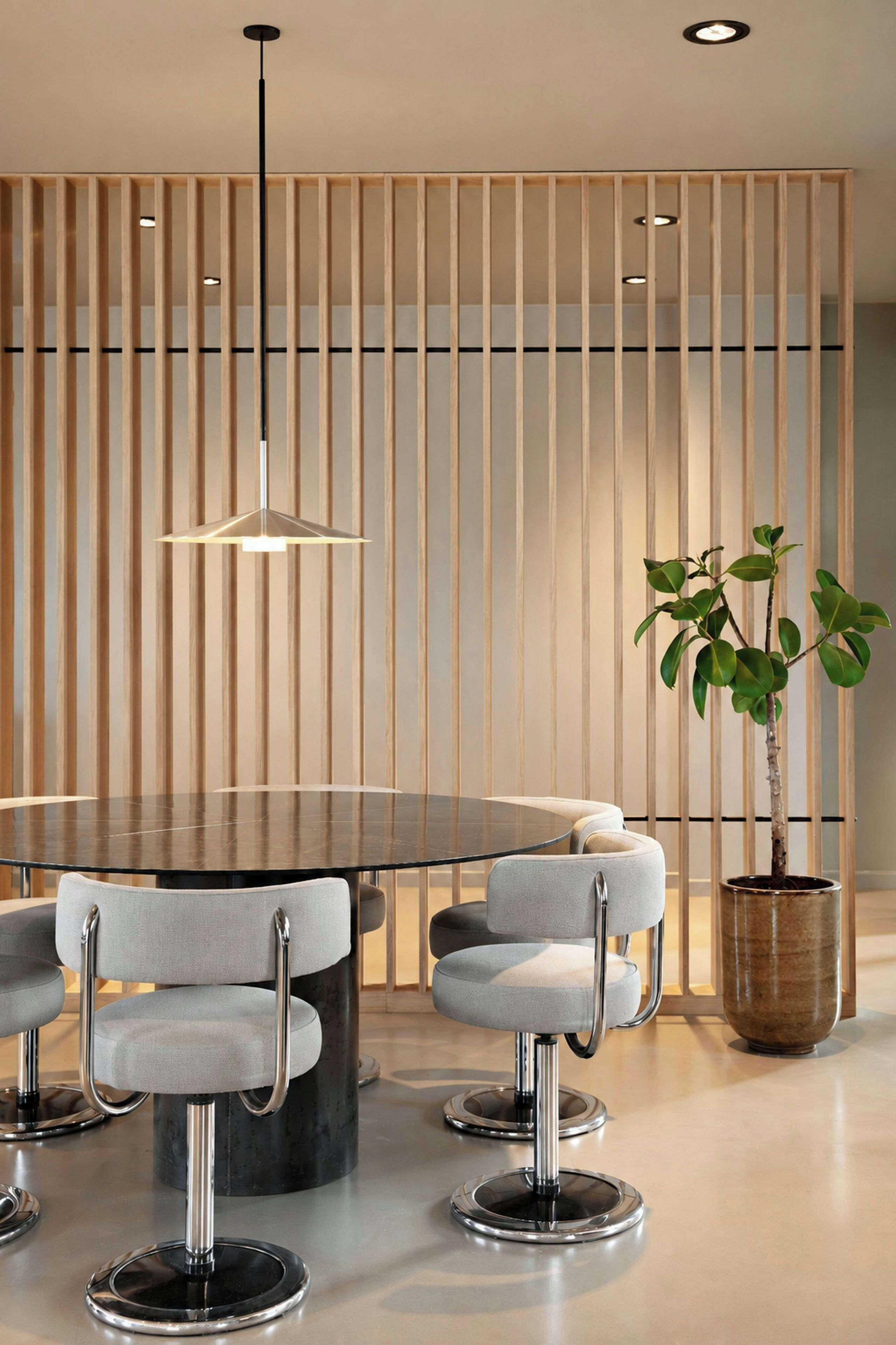 A modern dining area with a round table, grey upholstered chairs, a hanging light fixture, and a potted plant.