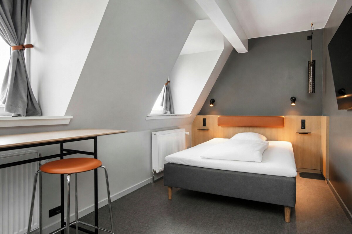 A modern bedroom with a sloped ceiling, a bed with white bedding, a desk with a stool, and a radiator.