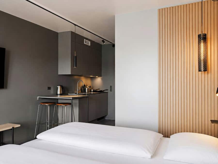 Un dormitorio moderno con una cama, una kitchenette pequeña y un televisor montado en la pared.