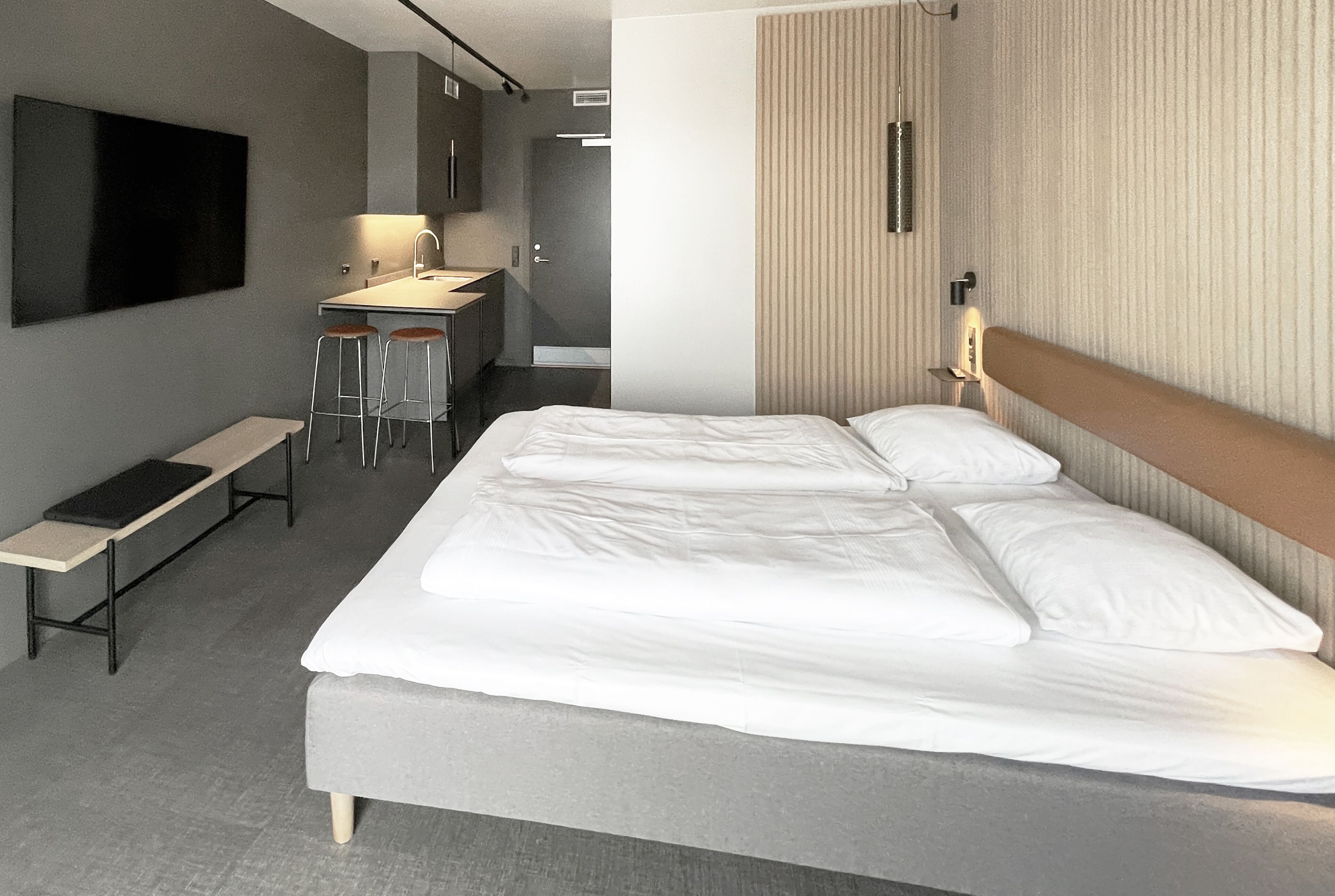 A modern hotel room with a large bed, a desk with stools, a bench, and a TV mounted on the wall.