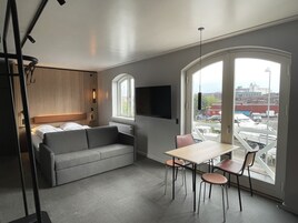 Una habitación moderna con un sofá gris, una pequeña mesa con sillas, un televisor montado en la pared y un ventanal con vistas a edificios.