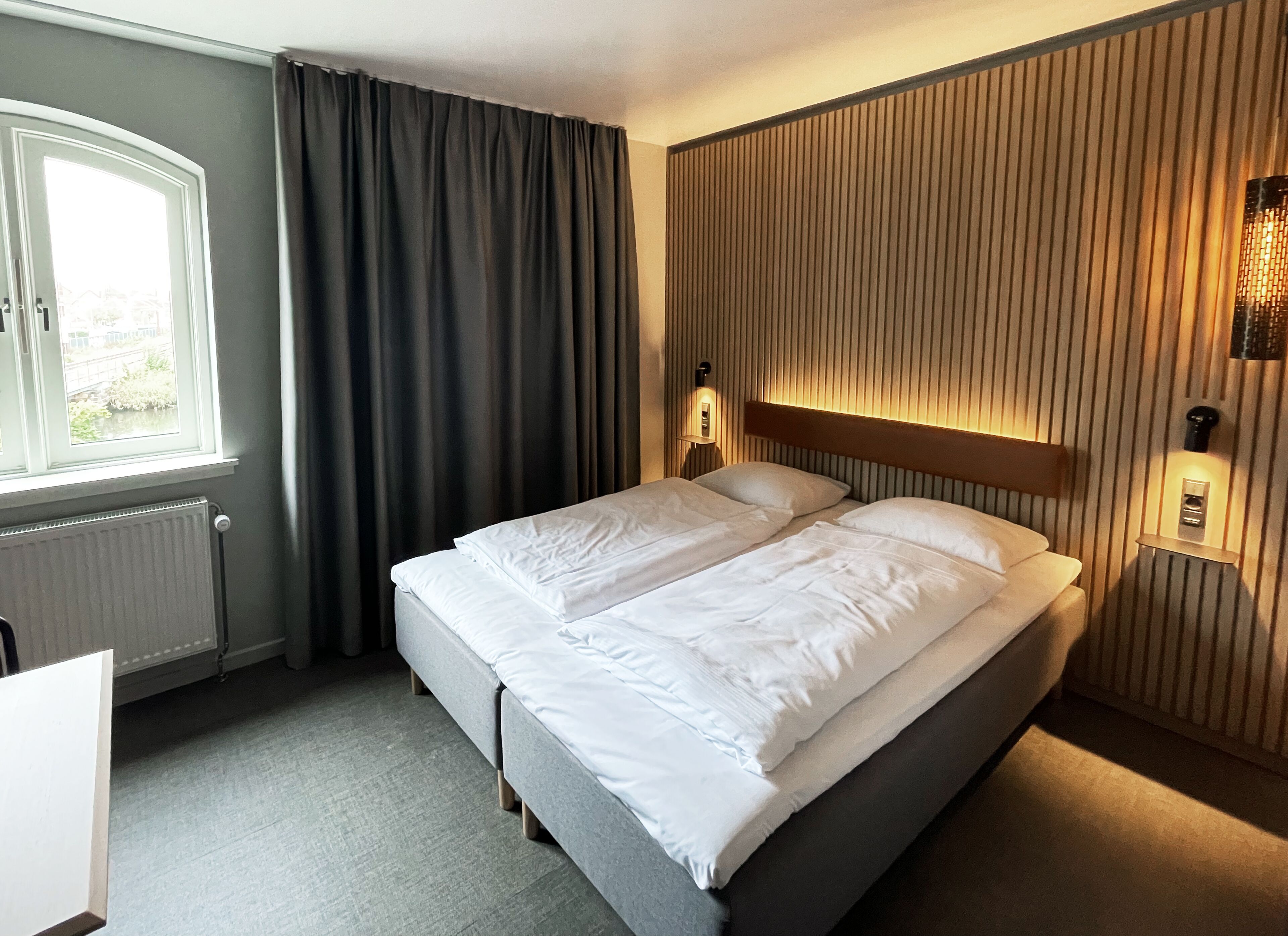 A hotel room with a bed, a window with curtains, a radiator, and a wall-mounted light fixture.