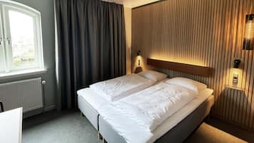 A hotel room with a bed, a window with curtains, a radiator, and a wall-mounted light fixture.