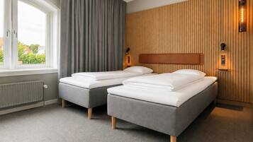 A hotel room with two beds, a large window with curtains, a radiator, and wooden paneling on the wall.