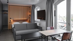 A modern hotel room with a sofa bed, a dining table with chairs, a TV mounted on the wall, and a large window offering a city view. - Four Points Flex by Sheraton Køge (Koge)