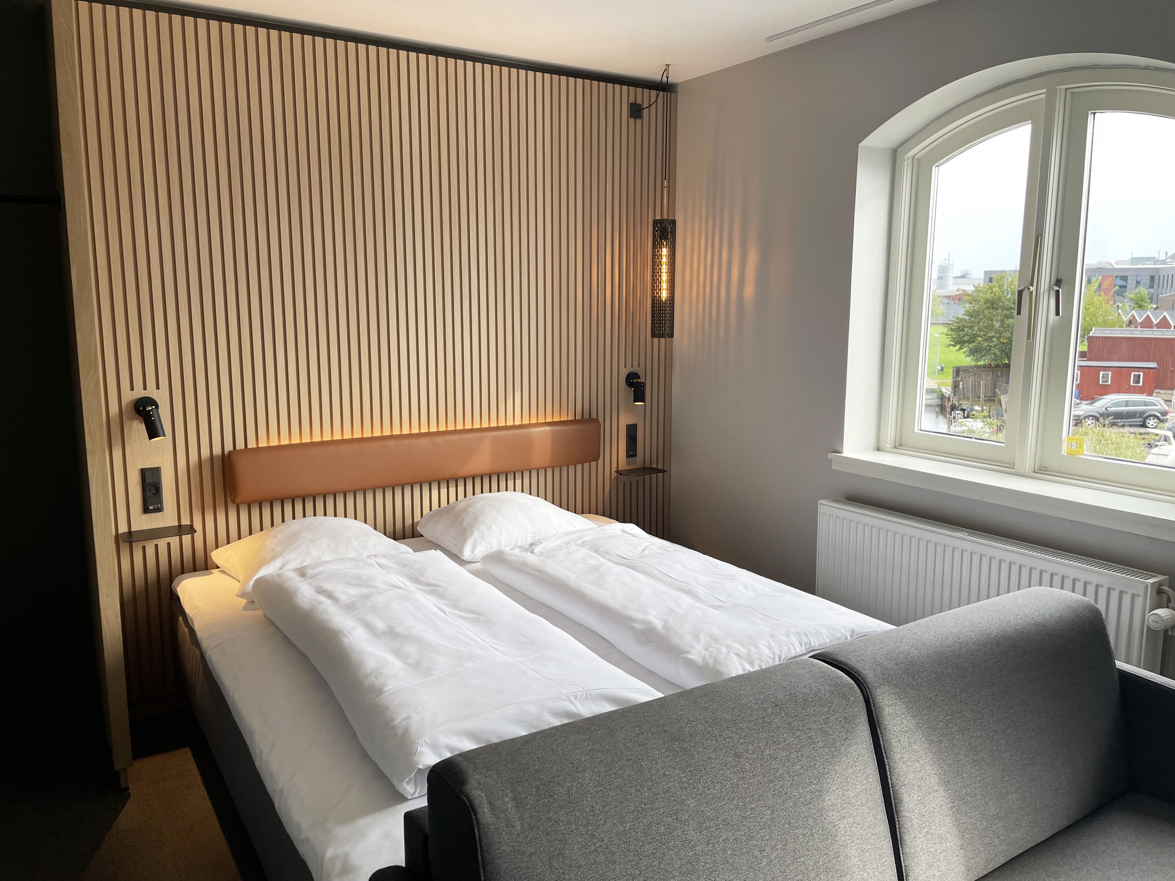 A hotel room with a bed, a bench, a window with a view, and a wall with vertical lines.