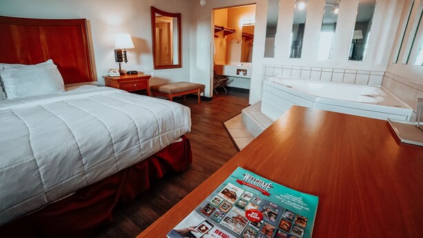 A hotel room with a bed, a wooden desk, a magazine on the desk, a chair, a small table, a mirror, and a bathroom visible through an open door. - Lamplighter Inn & Suites - North (Springfield)
