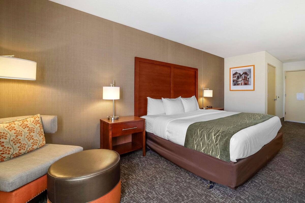 Ein Hotelzimmer mit einem großen Bett, einem Nachttisch, einer Lampe, einem Sofa und einem kleinen Hocker.