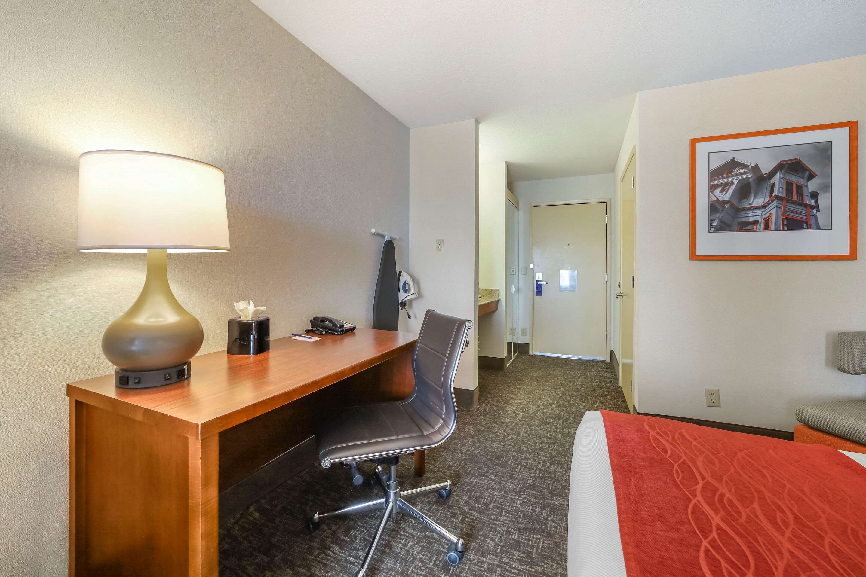 Une chambre d’hôtel avec un bureau en bois, une chaise, un lit recouvert d’une couverture rouge, une lampe et un tableau encadré accroché au mur.