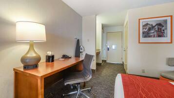 Une chambre d’hôtel avec un bureau en bois, une chaise, un lit recouvert d’une couverture rouge, une lampe et un tableau encadré accroché au mur.