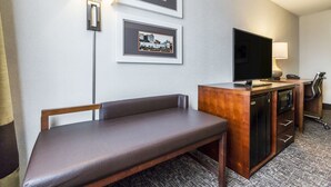 Habitación de hotel con un banco, televisión, escritorio con lámpara y cuadros en la pared.