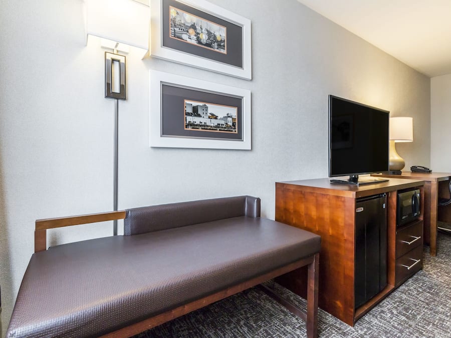 Habitación de hotel con un banco, televisión, escritorio con lámpara y cuadros en la pared.