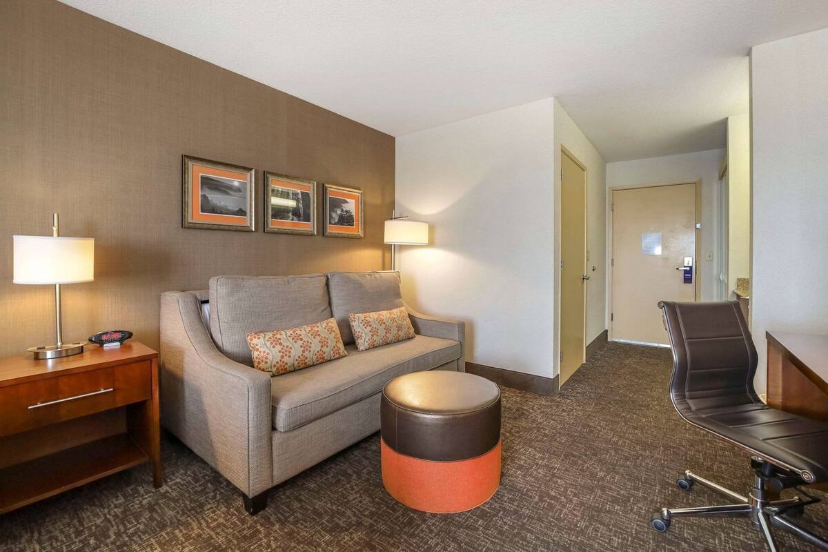 Ein Hotelzimmer mit einer Couch, einem kleinen runden Hocker, einem Schreibtisch mit Stuhl und gerahmten Bildern an der Wand.