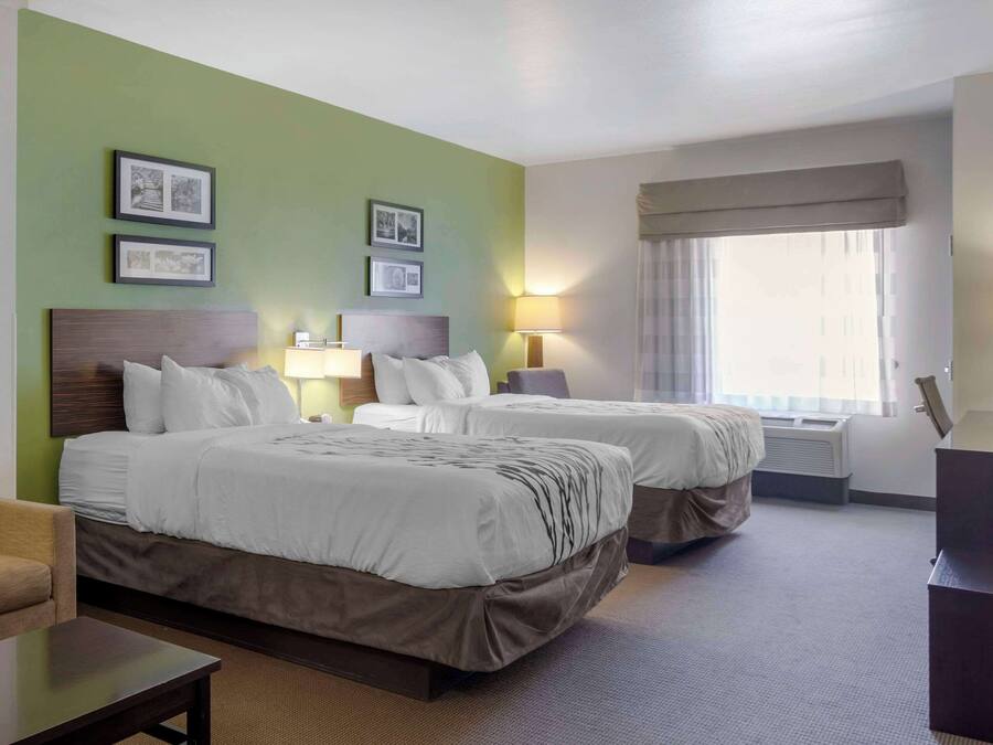 Ett hotellrum med två sängar, ett skrivbord och en stol. På väggen hänger inramade tavlor, det finns ett fönster med gardiner och en luftkonditioneringsanläggning.
