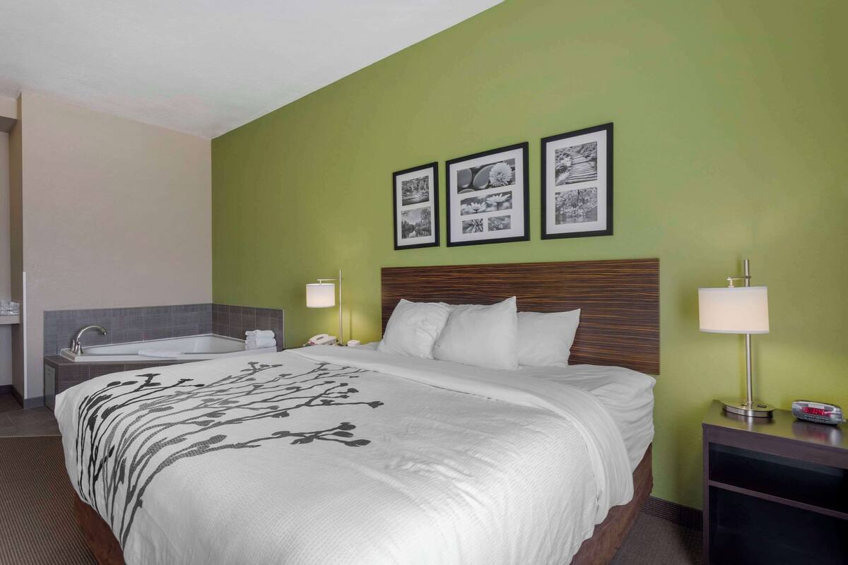A hotel room with a large bed, a bedside table with a lamp, a nightstand, a bathroom with a sink and mirror, and three framed pictures on the wall.