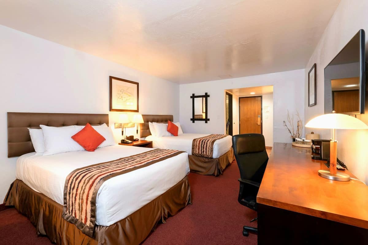 A hotel room with two beds, a desk, a chair, a lamp, and a door leading to another room.