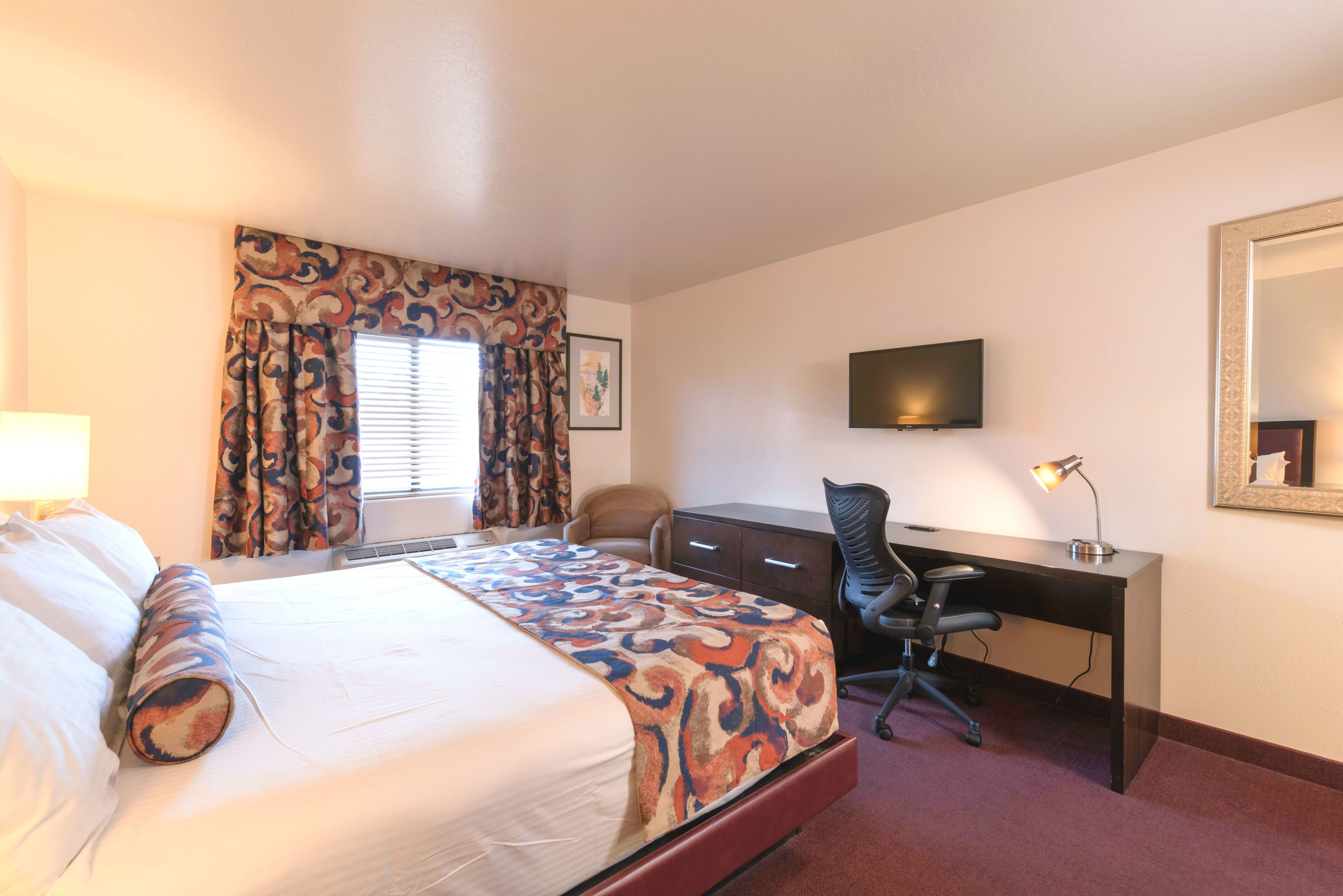 A hotel room with a bed, a desk, a chair, a TV, and a mirror.
