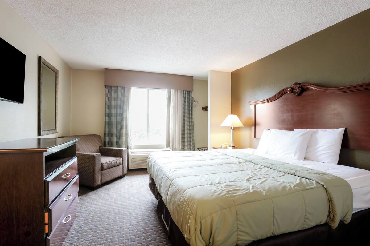 Une chambre d’hôtel avec un lit, une chaise, un bureau, une lampe et une fenêtre avec des rideaux.